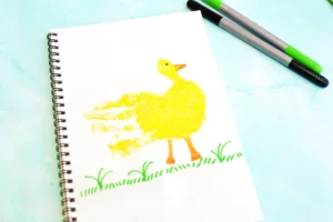 Spiral-bound notebook on a blue surface with a yellow duck painting; two markers lie diagonally nearby (green and gray/black) for this handprint animal craft
