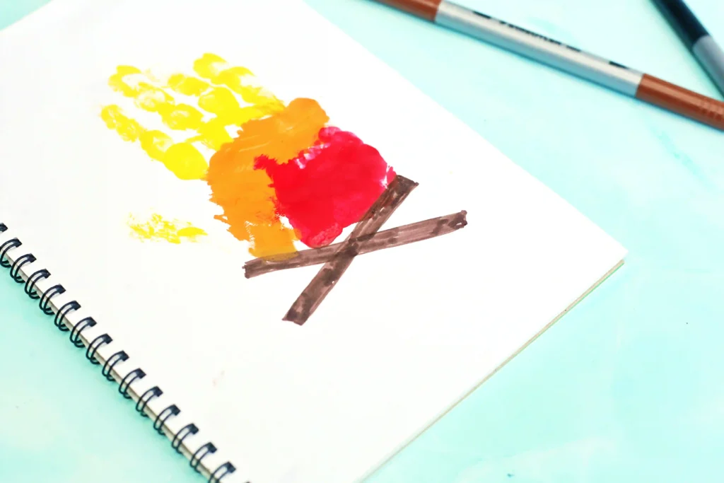 Open spiral-bound notebook on a light blue surface with yellow, orange and red handprint, and brown logs underneath.