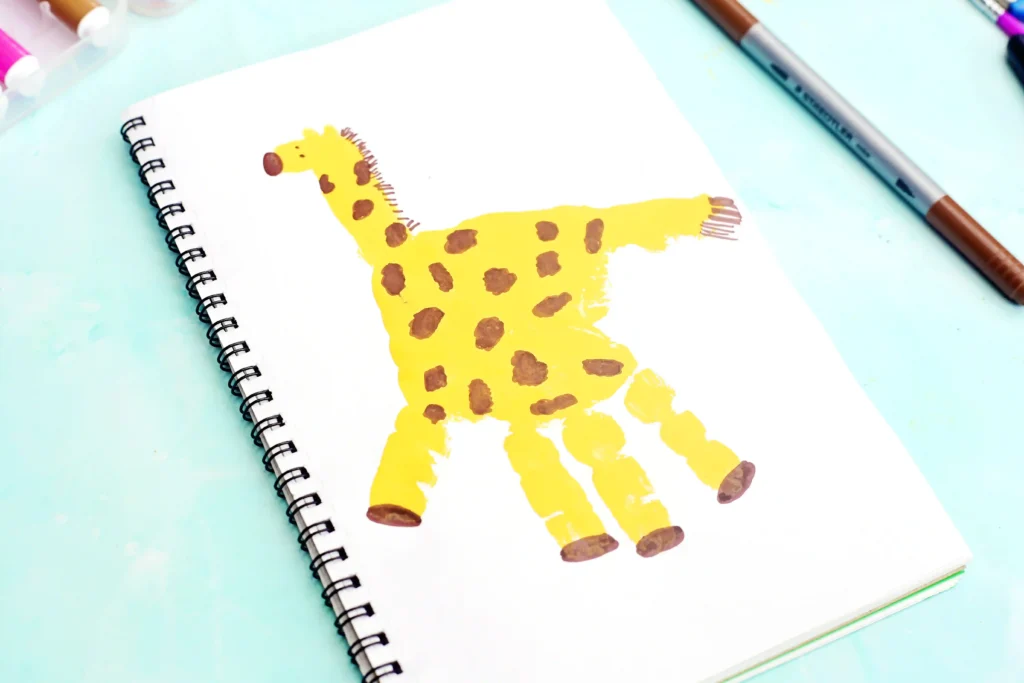 Notebook page with a bright yellow giraffe handprint and brown spots, surrounded by pastel art supplies on a light blue surface.
