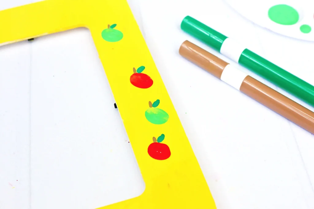 Diagonal view of a bright yellow photo frame with red and green fingerprint apples on a white surface, accompanied by green and brown markers nearby.