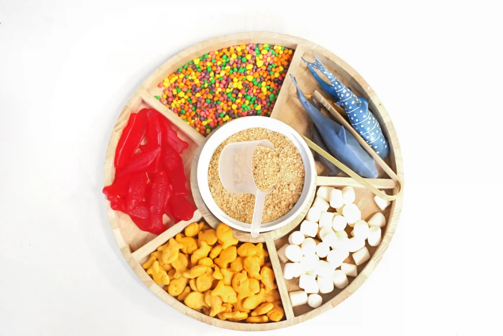a taste-safe sensory bin that is a Round wooden circular tray with separated sections holding colorful candies, red gummies, granola, white marshmallows, and blue toy sharks.