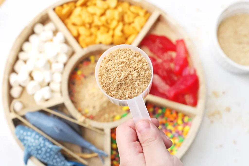 Close-up of a scoop filled with crushed graham crackers held over a tray with different candies and bowls in the blurred background.