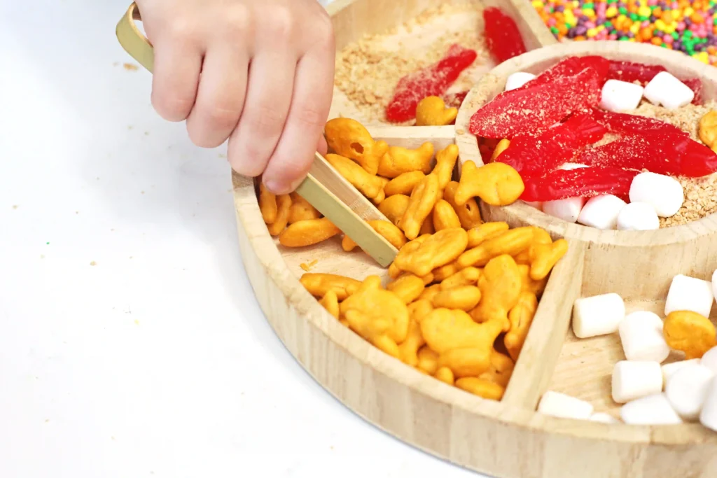 Hand scooping a wooden tray of mixed snacks: orange crackers, red gummy strips, marshmallows, and colorful sprinkles.