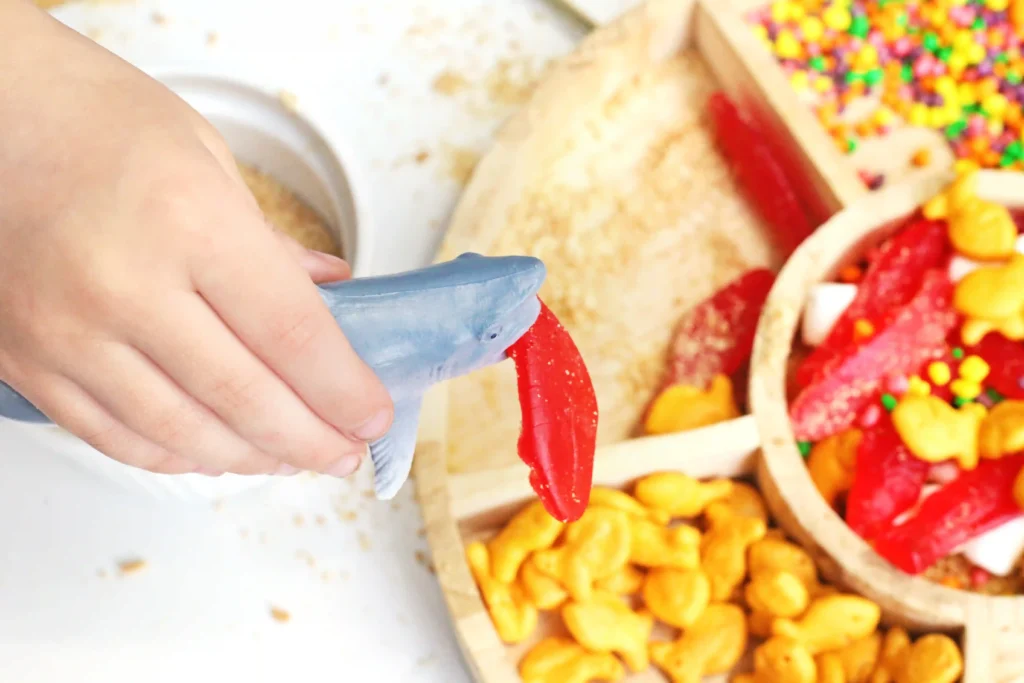 Close-up of a kid holding a toy shark with a red swedish fish in the shark's mouth over a tray of colorful sprinkles and snacks in the background.