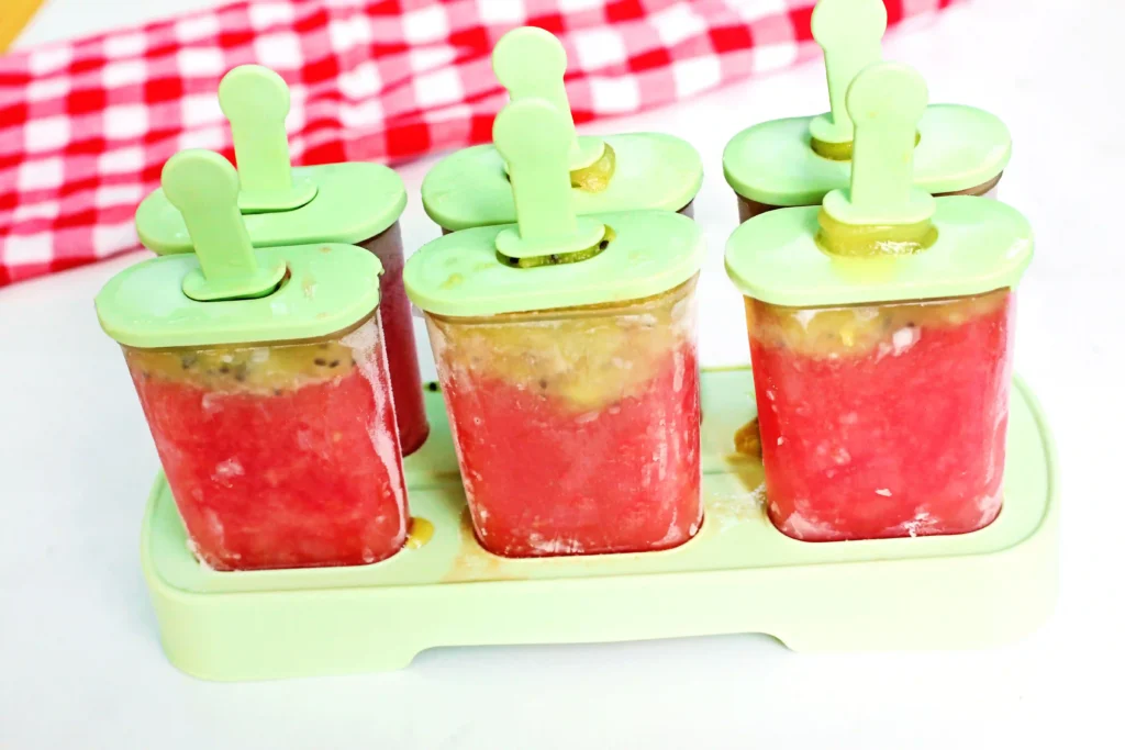 Watermelon Kiwi Popsicles - Cultivating Brilliant Minds Pastel green ice pop mold tray holding red fruit popsicles with green lids, set against a white surface and red checkered background.