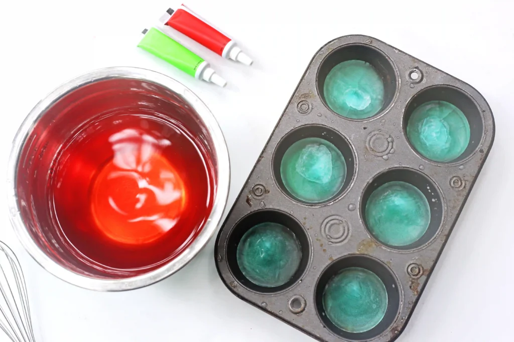 supplies for this sensory activity - A metal muffin tray with teal frozen spheres next to a glass bowl of bright red liquid and two squeeze tubes on a white surface, suggesting food coloring prep.