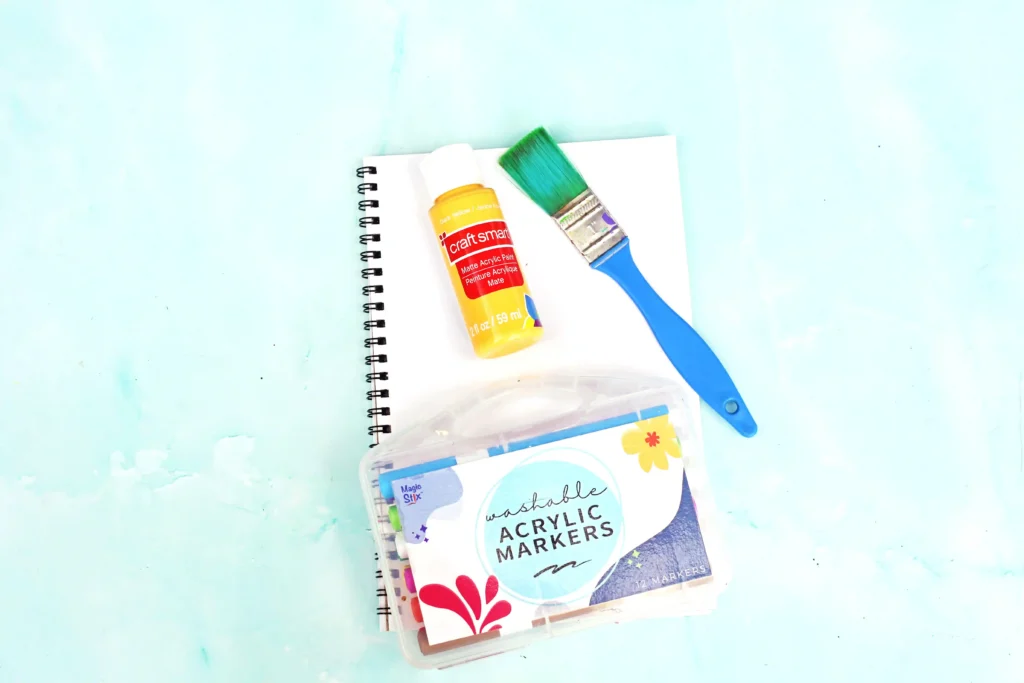 supplies for this animal craft for kids: Open spiral notebook with a yellow Craft Smart acrylic paint bottle and a green paintbrush on a pale blue surface.