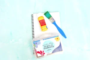 supplies for this animal craft for kids: Open spiral notebook with a yellow Craft Smart acrylic paint bottle and a green paintbrush on a pale blue surface.
