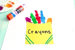 Rainbow-colored handprint art on yellow card, with crayons and glue stick nearby for a kids' craft.