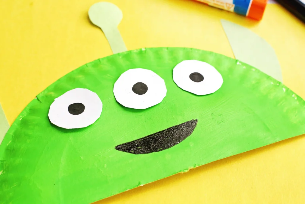 a Toy Story Alien Paper Plate Craft that is a paper plate cut in half, painted green, and decorated to look like the alien from toy story for a toy story birthday party