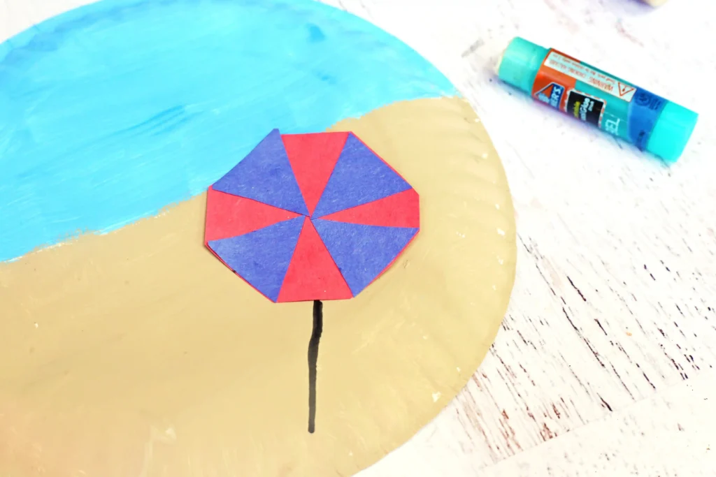 Close-up of a small red-and-blue umbrella on a beige circular plate, with a turquoise glue stick in the background.