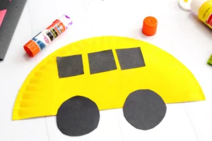 Craft project: yellow semicircle paper plate with black paper shapes (circles and rectangles) glued on a white surface to look like a school bus