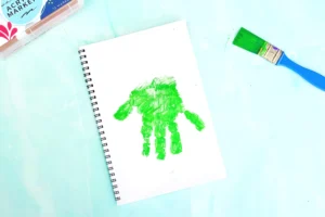 Notebook with a vivid green handprint stamp on its page, surrounded by green paint on a turquoise workspace with a brush nearby for this letter f alphabet craft for kids