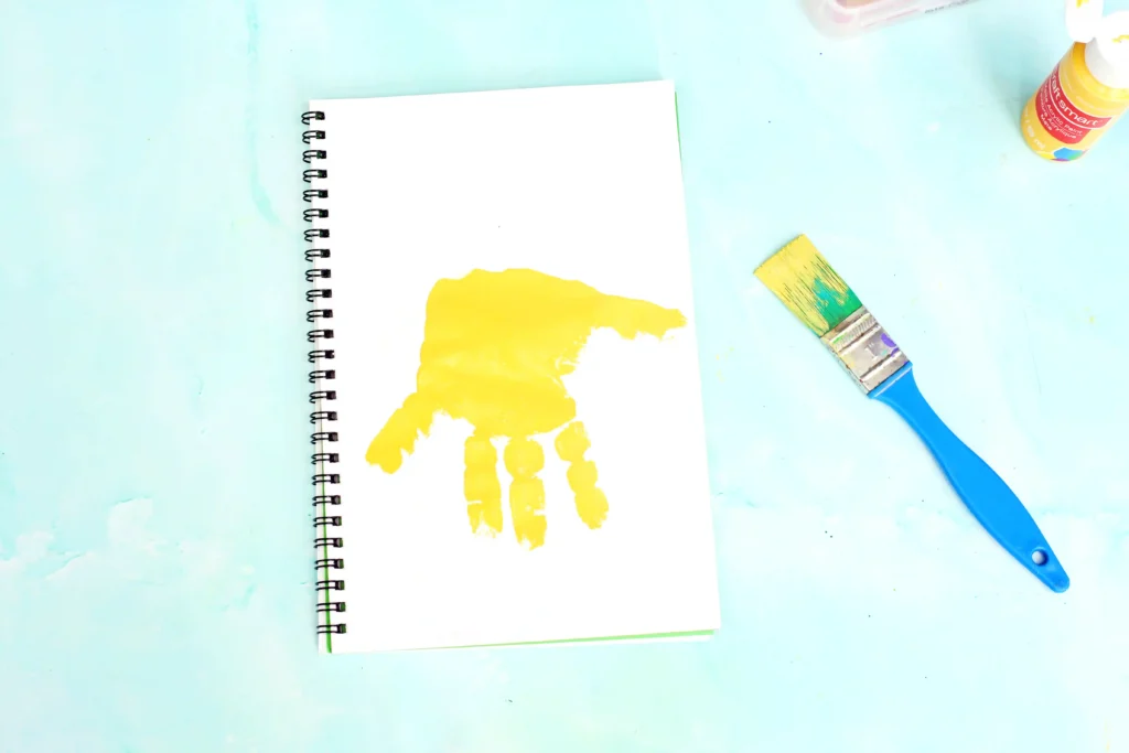 Notebook with a bright yellow handprint on its page, plus a blue-handled paintbrush and yellow paint bottle on a pastel blue surface for this Alphabet craft for kids