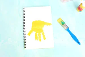 Notebook with a bright yellow handprint on its page, plus a blue-handled paintbrush and yellow paint bottle on a pastel blue surface for this Alphabet craft for kids