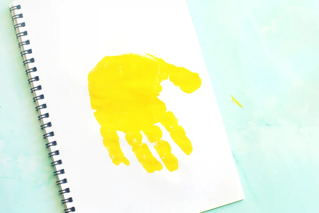 Open spiral notebook with a bright yellow handprint on a blank page, resting on a pale blue surface for this letter J handprint craft