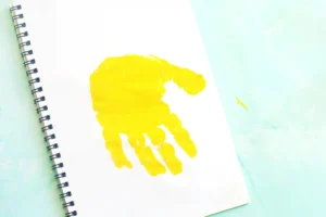 Open spiral notebook with a bright yellow handprint on a blank page, resting on a pale blue surface for this letter J handprint craft