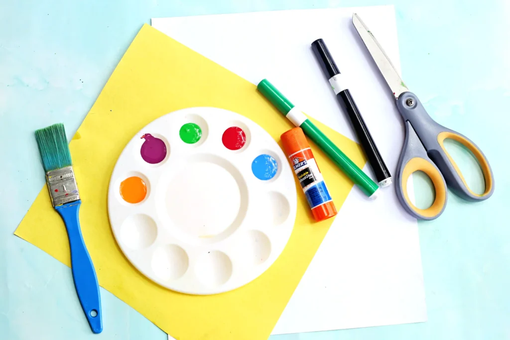 Supplies for this crayon craft- paint palette with colored paints, glue stick, markers, scissors, and a brush on a blue background.
