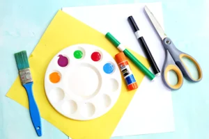 Supplies for this crayon craft- paint palette with colored paints, glue stick, markers, scissors, and a brush on a blue background.