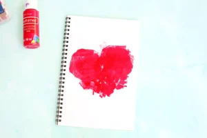 Notebook page with a large red paint smear and a red craft paint bottle beside it on a light blue surface.