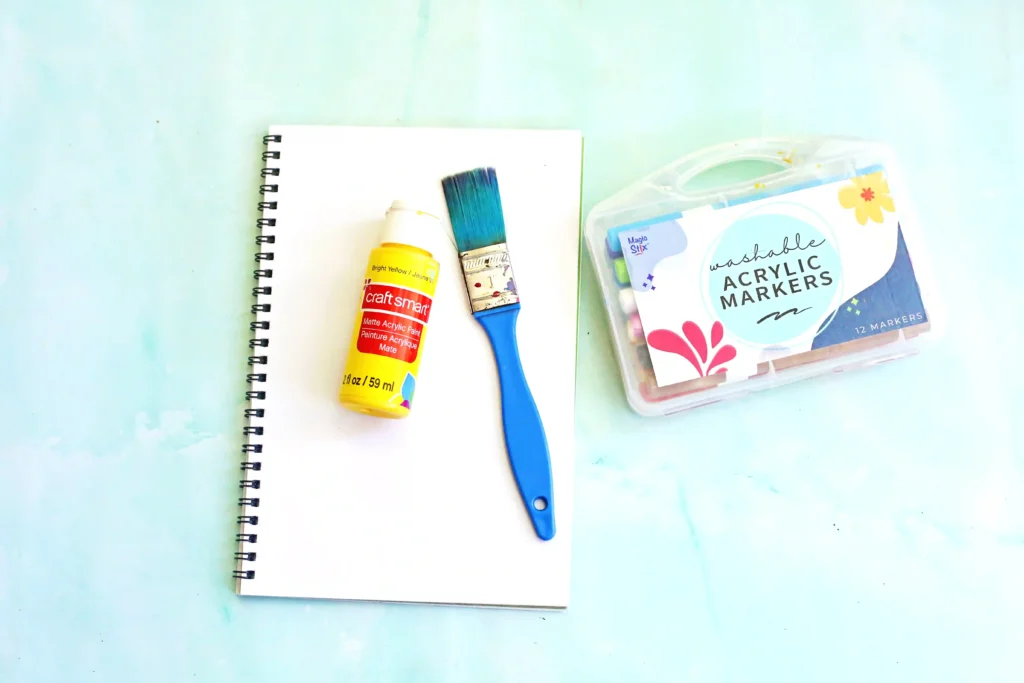 supplies for this j is for jaguar handprint craft: Spiral notebook with white pages alongside a yellow craft paint bottle, blue paintbrush, and a box of washable acrylic markers on a light surface.