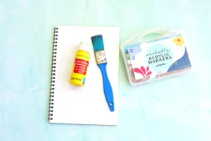 supplies for this j is for jaguar handprint craft: Spiral notebook with white pages alongside a yellow craft paint bottle, blue paintbrush, and a box of washable acrylic markers on a light surface.