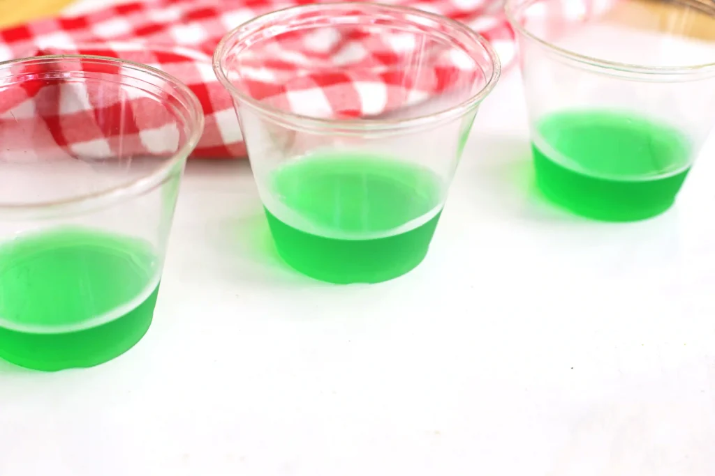 Three clear plastic cups partially filled with bright green jello on a white surface, with a red-and-white checkered cloth in the background.