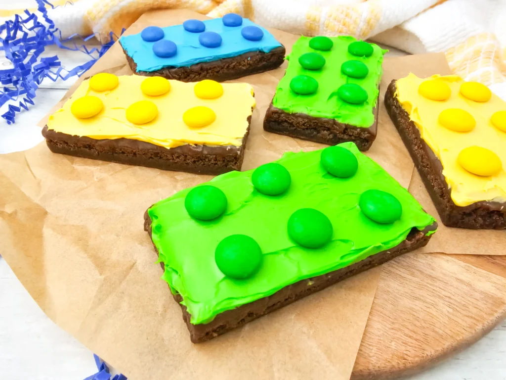 Assorted square brownies with bright blue, yellow, and green icing and matching round candies on parchment.