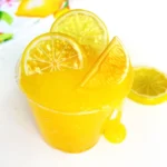 Tall glass of a lemon slime recipe with lemon slices and a lemon wedge, set against a bright background with citrus decor.