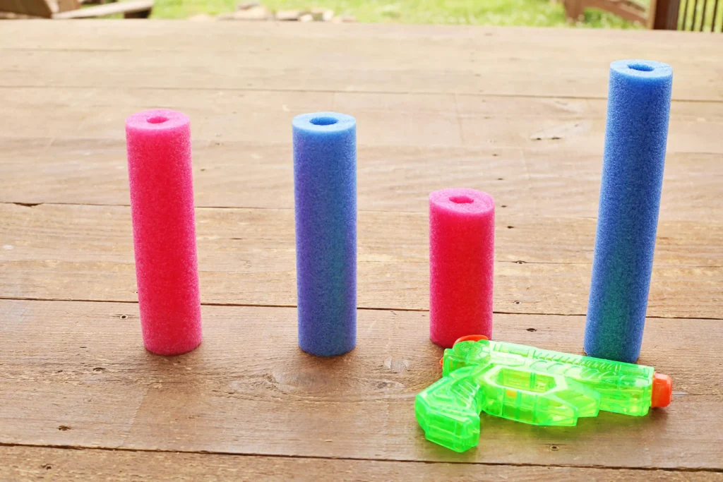 Outdoor Water Play Activity - Cultivating Brilliant Minds pink and blue pool noodles next to a green squirt gun