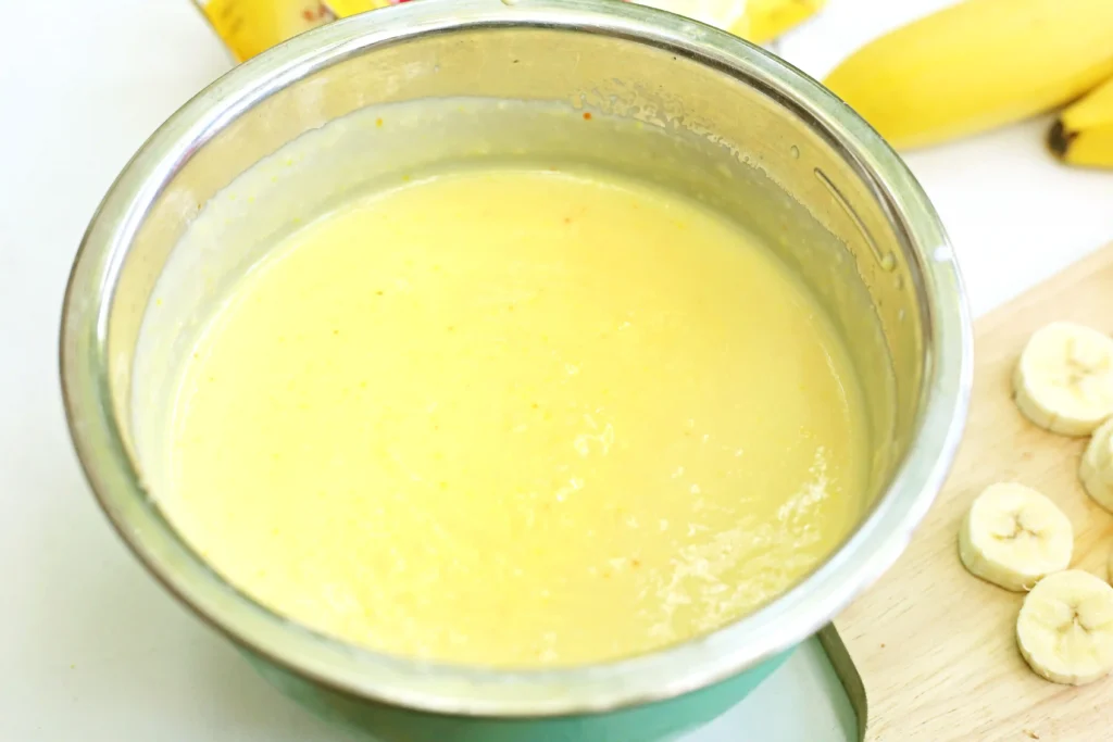 Banana pudding batter in a large metal mixing bowl with banana slices on a wooden board nearby.