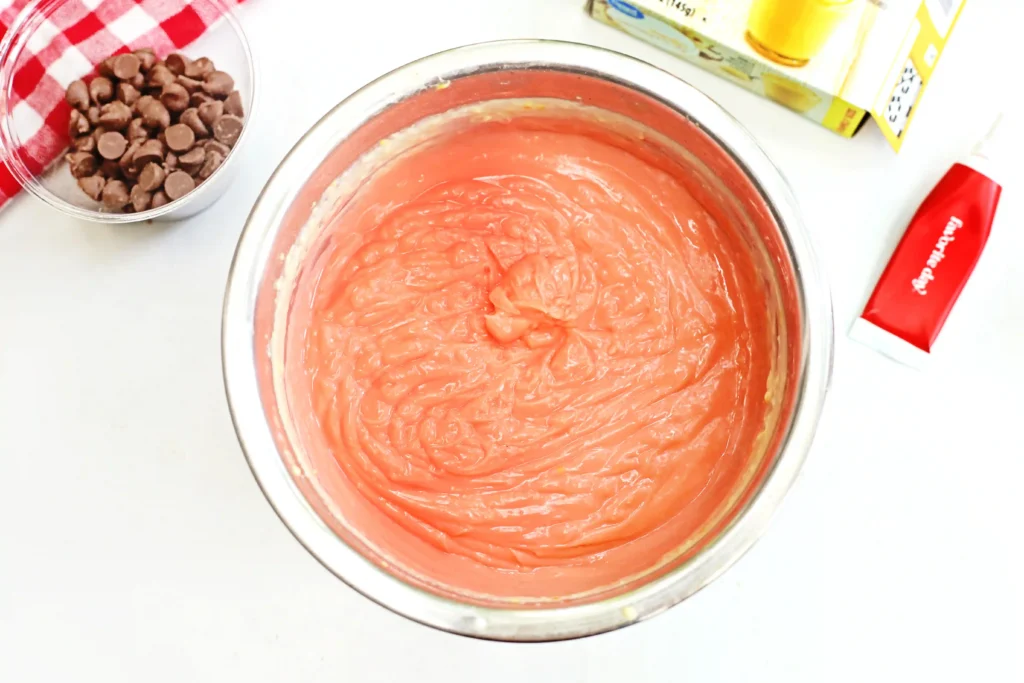 Pink dyed vanilla pudding in a glass bowl with a cup of chocolate chips nearby and a red spatula on the counter.