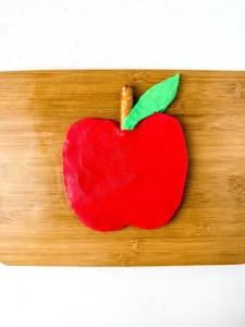Red apple-shaped buttercream on a wooden cutting board, with a small brown stem and a green leaf protruding from the top edge