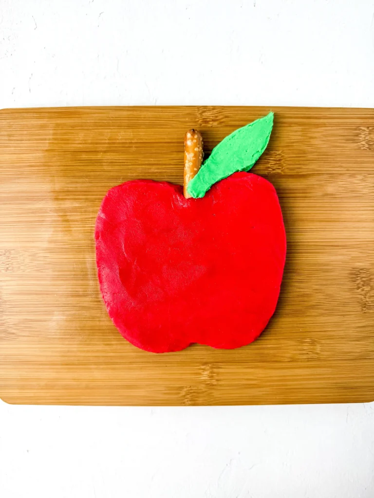 Red apple-shaped buttercream on a wooden cutting board, with a small brown stem and a green leaf protruding from the top edge