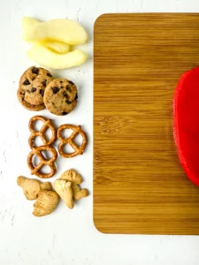Assorted cookies, pretzels, and apple slices on a white surface beside a wooden cutting board.