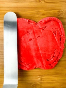 Red buttercream shaped into an apple with a stainless steel spatula resting on a wooden surface on top of a cutting board for this party dessert board