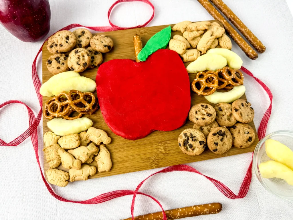 an Apple Buttercream Board that has a buttercream apple surrounded by apple slices, cookies, and pretzels on a wooden baord