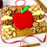 an Apple Buttercream Board that has a buttercream apple surrounded by apple slices, cookies, and pretzels on a wooden baord