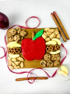 an Apple Buttercream Board that has a buttercream apple surrounded by apple slices, cookies, and pretzels on a wooden baord