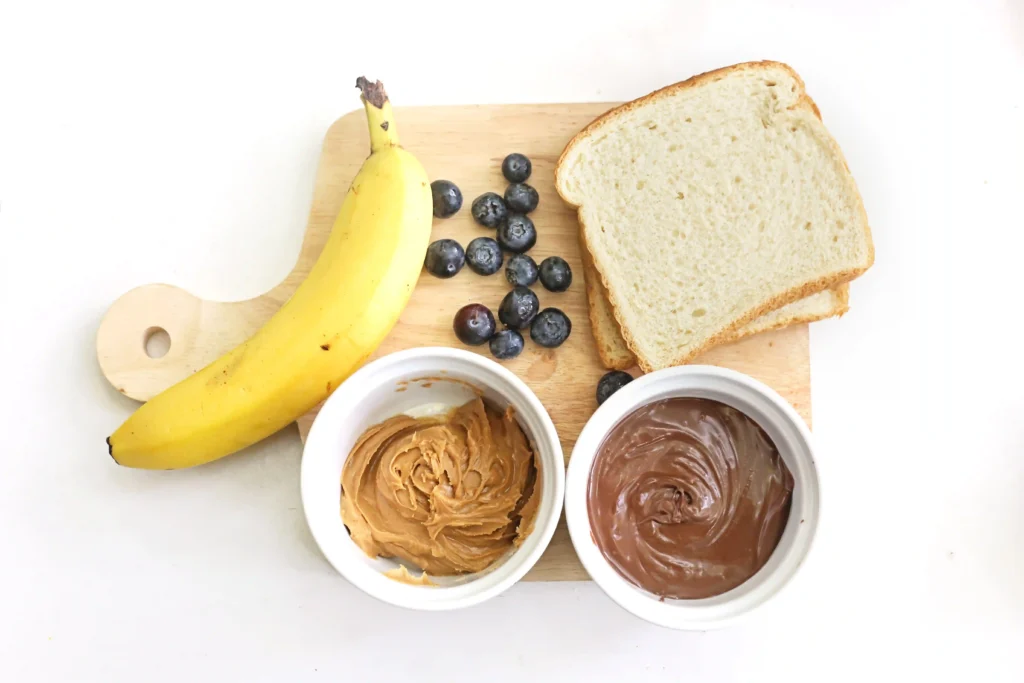 ingredients for this easy breakfast idea for kids- Banana and blueberries on a wooden cutting board with two slices of bread and bowls of peanut butter and chocolate spread below.
