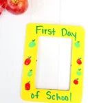 a First Day of School Photo Frame that is painted yellow with fingerprint apples and words that read "first day of school"