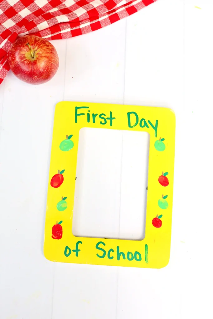 a First Day of School Photo Frame that is painted yellow with fingerprint apples and words that read "first day of school"