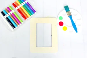 supplies for this back to school craft: Assorted colored markers in a case beside a paint palette an unpainted photo frame, and blue brush on a white surface, ready for art.