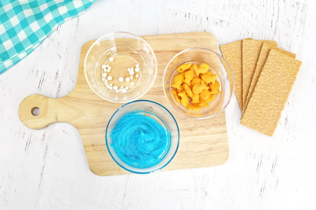 Ocean Graham Crackers - Cultivating Brilliant Minds supplies for this easy summer snack - Top-down view of a wooden board with bowls of white candies and orange snacks, a blue beverage, crackers, and a teal checkered cloth in the corner.