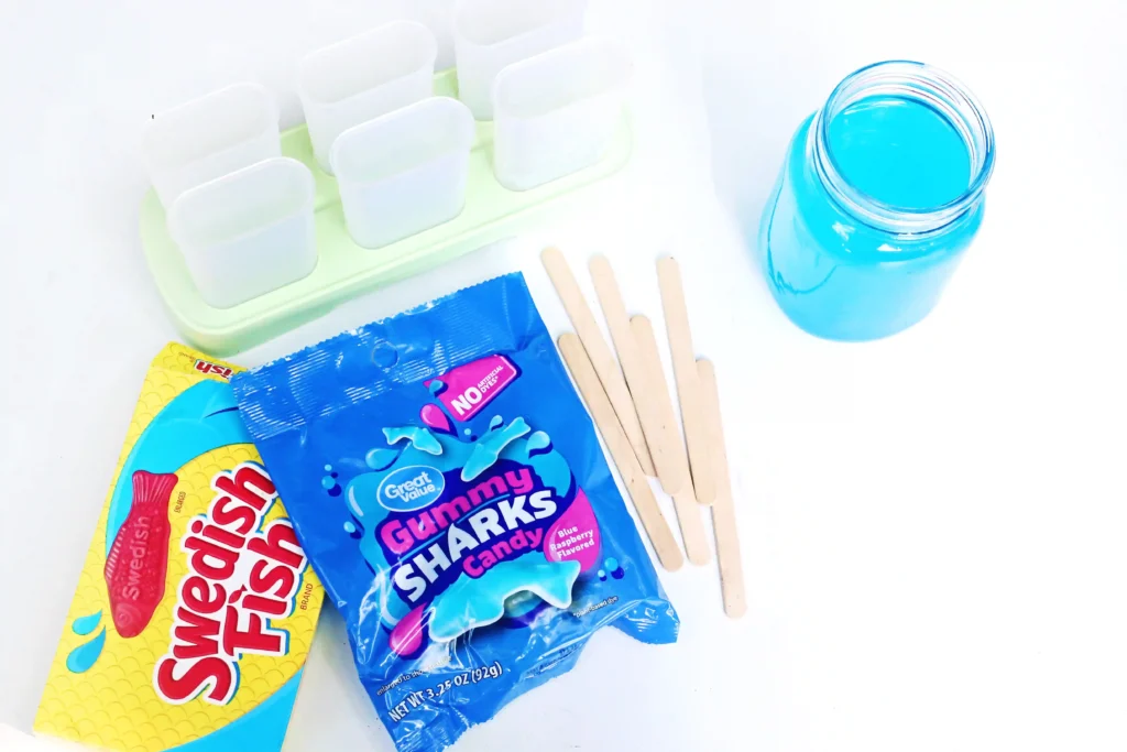 ingredients for these popsicle ideas - Assortment of candy and snack items on a white surface: blue gummy sharks candy bag, yellow snack wrapper, wooden popsicle sticks, and a blue liquid in a jar nearby.