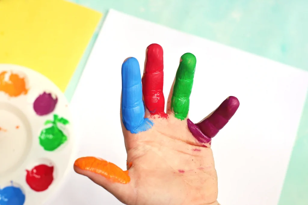 Child's hand with multicolored painted fingers (blue, red, green, purple) during finger painting activity; paint palette in background.