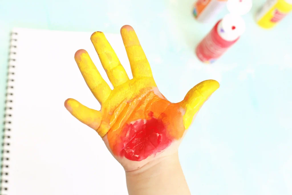 Hand covered in bright yellow and red paint, raised in front of a blurred art desk background with paint bottles and a notebook for this c is for campfire handprint craft