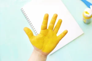 Hand raised with yellow paint on skin, over a notebook and paint bottle on a pale blue surface for this D is for duck handprint craft