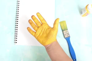 Overhead view of a painted hand in yellow-orange paint, with a paint bottle and blue scissors on a pale blue surface beside a spiral notebook for this giraffe craft