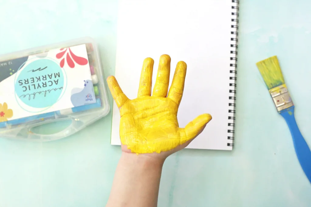 Hand with bright yellow paint on palm and fingers resting above a white notebook, with a paintbrush and art supplies nearby on a light blue surface for this handprint art